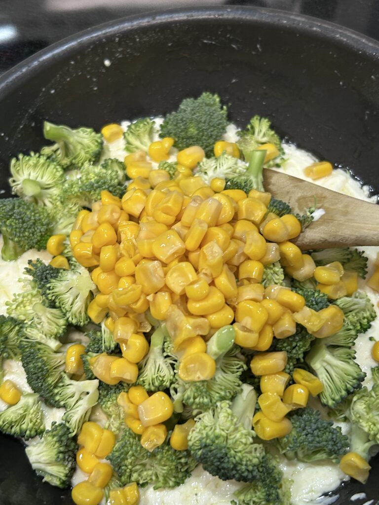 corn and broccoli florets with cream cheese  and chicken broth in a black saucepan.